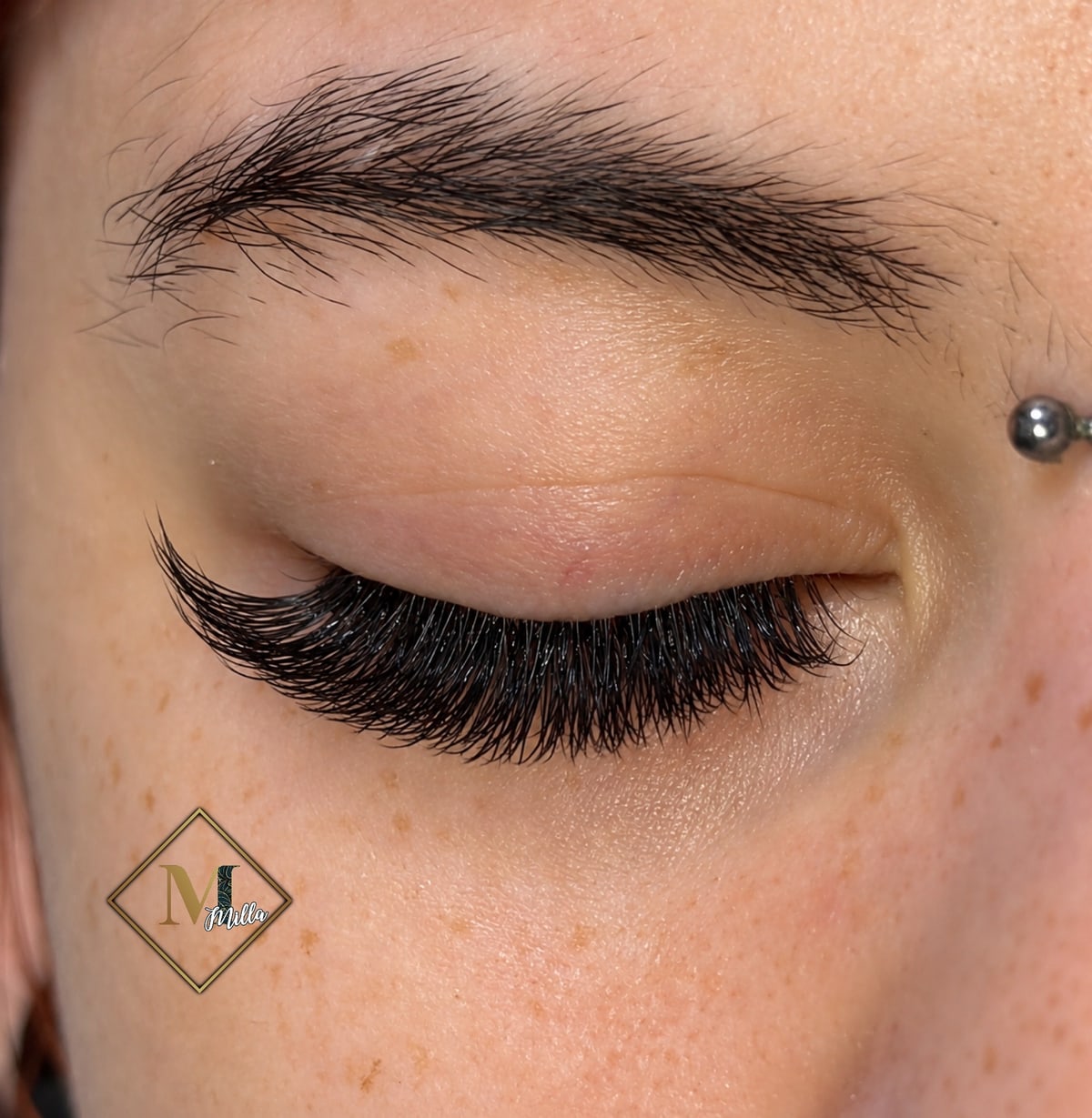 Közeli felvétel egy csukott szemről szeplős bőrön, hosszú, sűrű fekete épített szempillákkal, a szemöldök fölött egy kis piercinggel